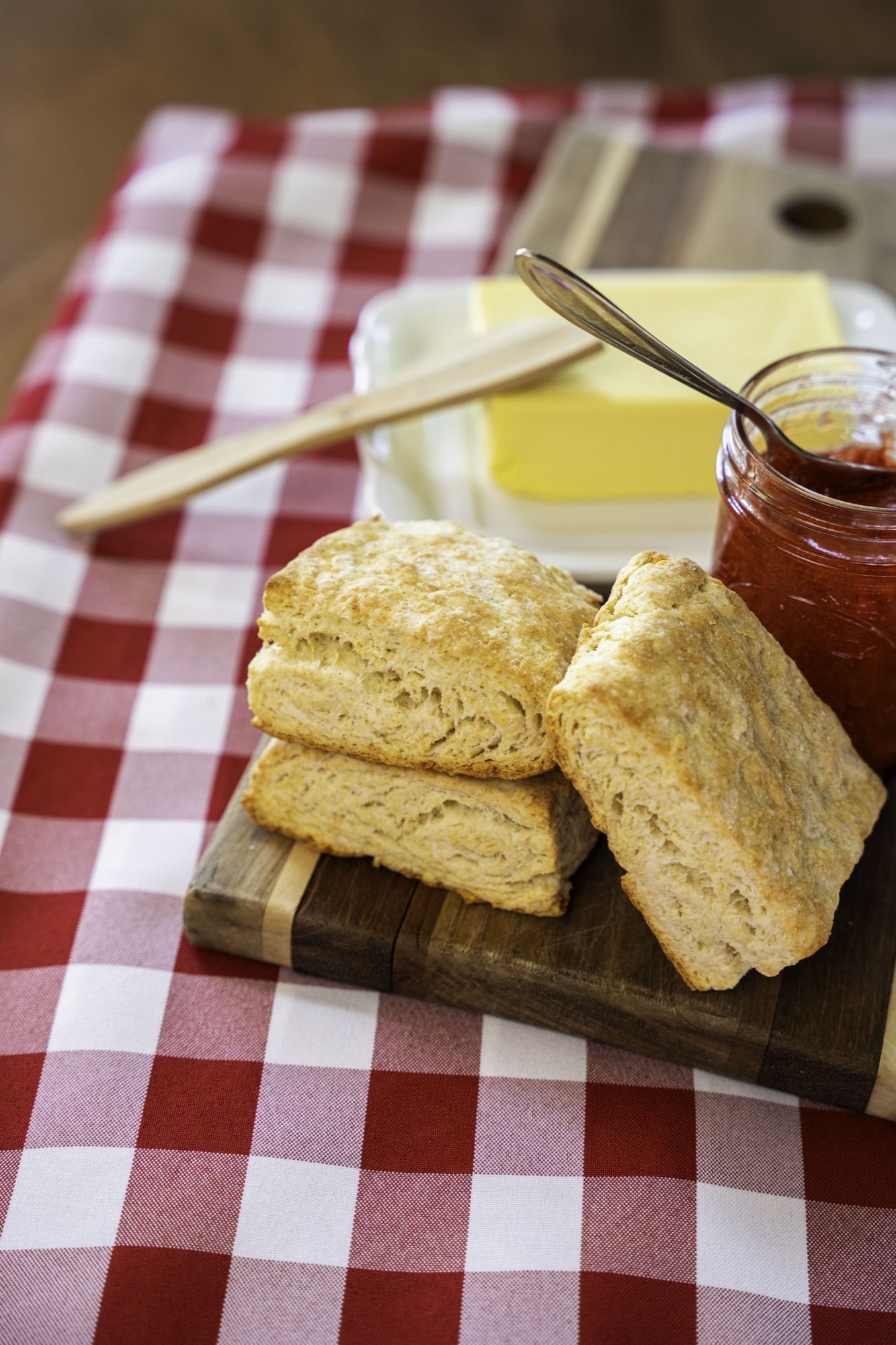 Flaky biscuits stacked on a wooden board with butter and a jar of strawberry jam.