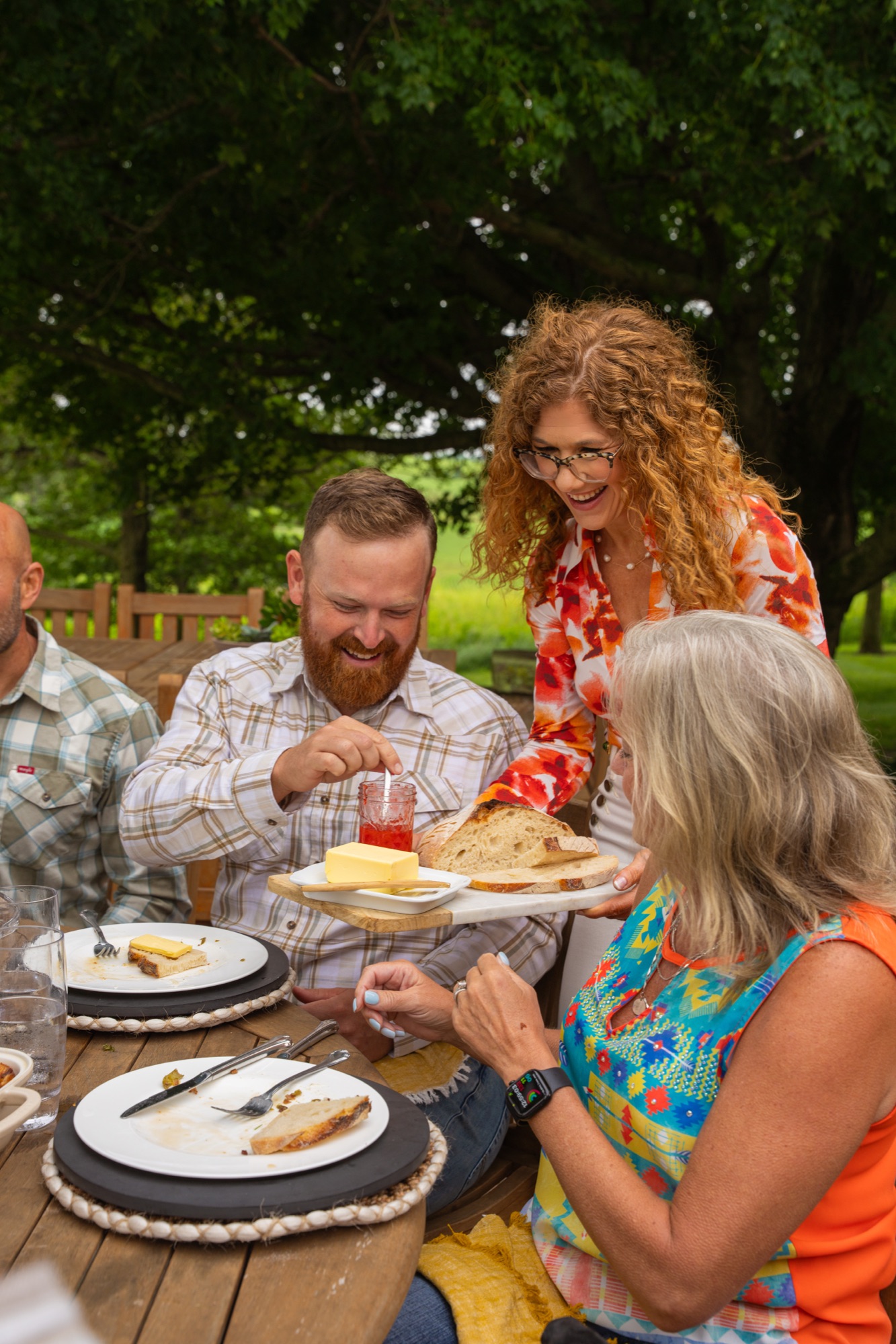 Sara passing a board with bread, butter, and jam to guests seated at an outdoor table.